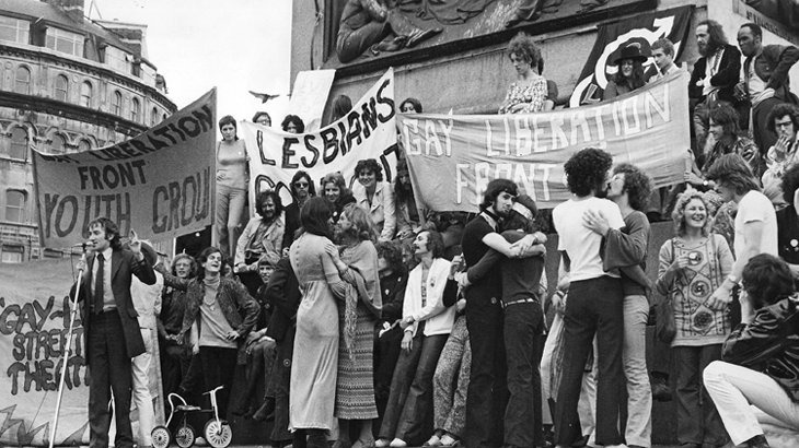 Alt: black and white photo of 1972 London Pride, the first Pride. There is a large group of people in Trafalgar Square holding banners saying 'Gay Liberation Front' and similar but somewhat obstructed sentiments on other banners. A pair of women and a pair of men are respectively making out while someone speaks from a microphone.